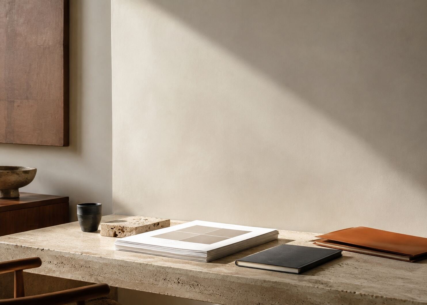 Quiet studio corner with blank layout proofs, a notebook, and a clay-orange folder on a limestone desk